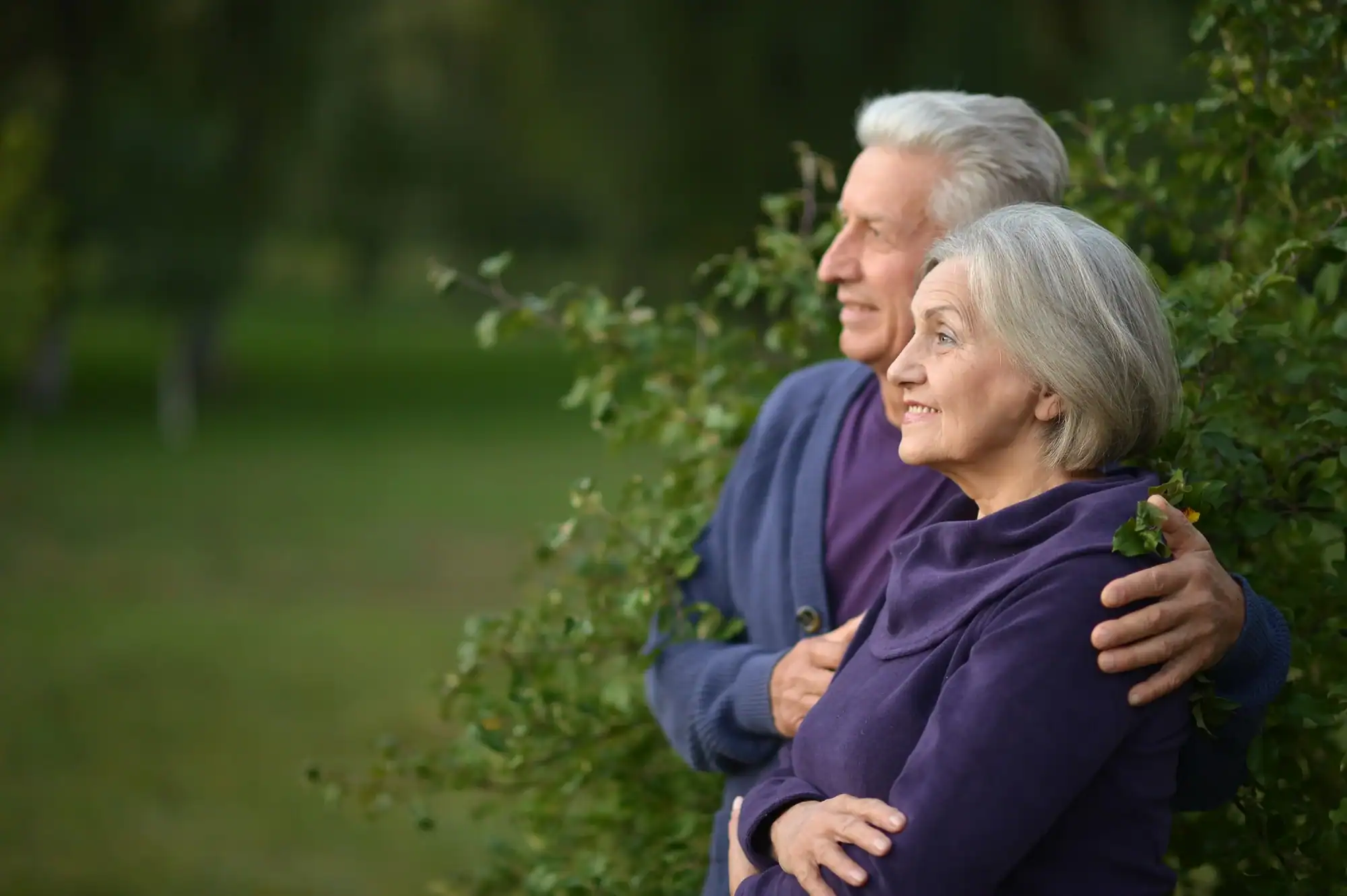 An elderly couple stands embracing, gazing contentedly into the distance
