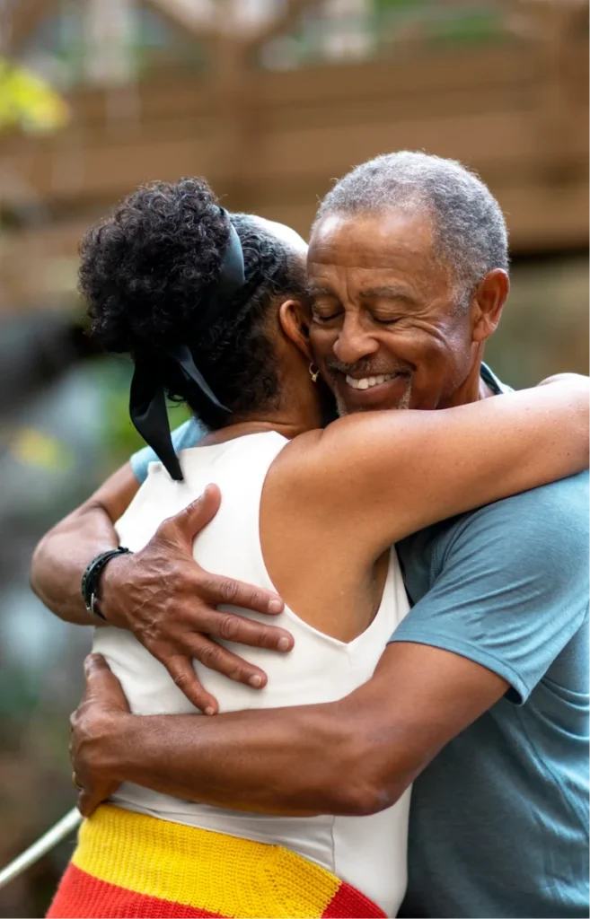 An elderly embraces warmly outdoors, smiling with eyes closed